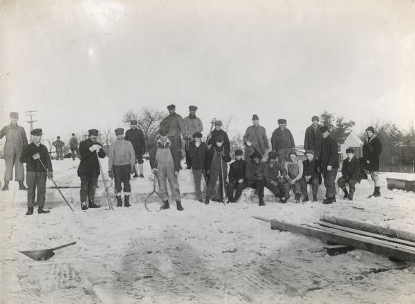 A large group of ice cutters posing for a portrait. They are holding a variety of implements and are sitting and standing on blocks of ice. Fence and buildings are in the background.