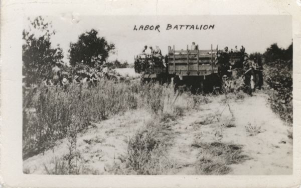 A group of Civilian Conservation Corps (CCC) men working in a field. On a rough road are three trucks. Two of the trucks have large groups of men in them. The third appears to be mostly empty, and a small group of men stand on the right near the truck. Another group of men sit and stand alongside the road in the brush on the left.