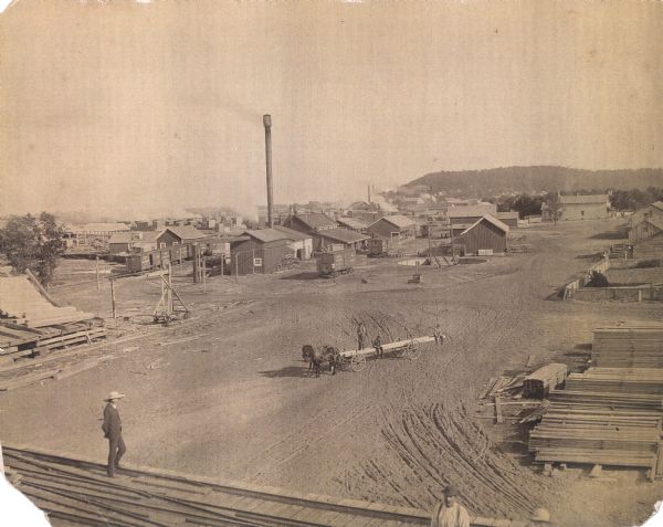 Elevated view of a lumber mill. Piles of milled wood are in the foreground and on the right, with more stacks in the far background behind numerous small buildings and a tall chimney. Working men are posed, one standing on a stack of wood on the left, and three men posing on a horse-drawn wagon. In the far background is a bluff.