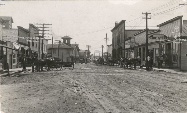 View down unpaved street, which has numerous horse-drawn vehicles parked along the curbs. People are on the street and sidewalk, with a larger group and a dog gathered in front of the shoe store on the corner on the right. The storefronts include O.A. Mattas and the J. Gross City Store.