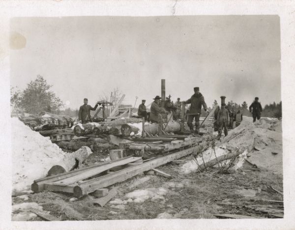 Group of men working outdoors in the snow. Large logs are stacked and ready to be cut. There are two steam or coal-powered machines in the background running a belt-driven saw.