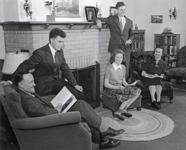 The Orland Loomis family relaxing at home. Mrs. Loomis holds a book, and leans slightly to look at her husband, who is also holding a book. A daughter sits on a stool in front of a fireplace holding a book. One son is standing and leaning on the mantlepiece, the other son is sitting on the arm of his father's chair.<p>Orland Loomis was elected Wisconsin governor in 1942 over the incumbent Republican, Julius P. Heil, but died before taking office.