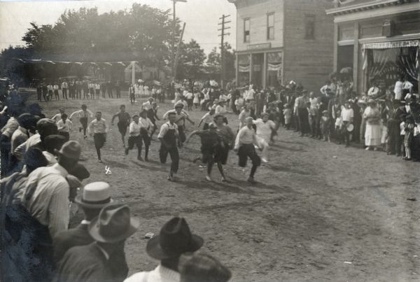 Slightly elevated view of a crowd of teenage boys racing down the street during the Hustler Harvest Festival. Both sides of the street are lined with crowds of people. In the background on the starting line is a row of men and boys. To their right are three girls wearing white dresses. An awning with a sign for the Hustler State Bank is on the right.