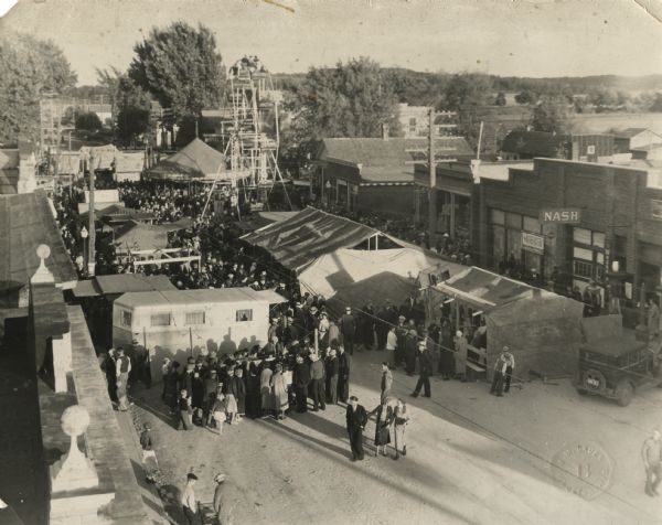 Elevated view of street lined with merchandise stalls, a merry-go-round and a Ferris wheel set up for the Harvest Festival. A large crowd of people is in line at each of the stalls. Across the street on the right is the Nash service station, and next to it is an ice cream shop.