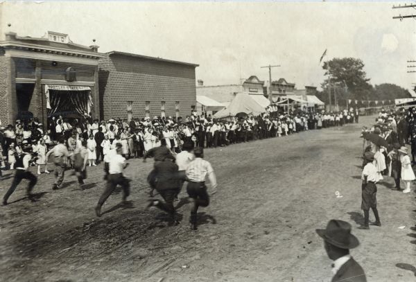 A group of boys is racing down Hustler's Main Street during the Harvest Festival. The finish line is in the distance at the far end of the street. Crowds stand and watch from the sidewalks. The Hustler State Bank is on across the street.