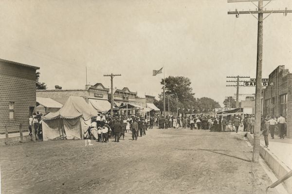 View down unpaved street towards a large crowd of men, women and children gathered around the stalls of the Hustler Harvest Festival. The festival is set up on Hustler's main street. On the left side are the A.J. Pitel General Merchandise store, the A.C. Schucht Hardware and Furniture Store. On the right side are the Nash Service Station and a garage. Somewhat obscured by a stall is a sign that says: "This car will be given away."