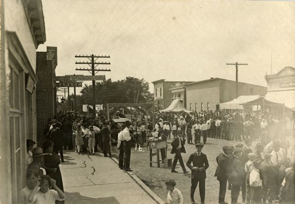 Slightly elevated view from sidewalk of a crowd of men, women and children gathered about the stalls at the Hustler Harvest Festival. On the left side three business names are visible: Nash Service Station, Martinson Brothers Shop, M.O. Rider Garage. Across the street is the Hustler State Bank, and the A.J. Pitel General Merchandise store.