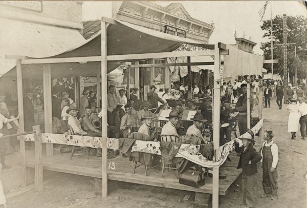 An African American military band poses for the camera during the Hustler Harvest Festival. They are sitting in an open sided structure with a canvas roof that extends into the street. In the background along the sidewalk is an International Harvester McCormick-Deering storefront. Further down the street is an American flag on a tall flagpole.
