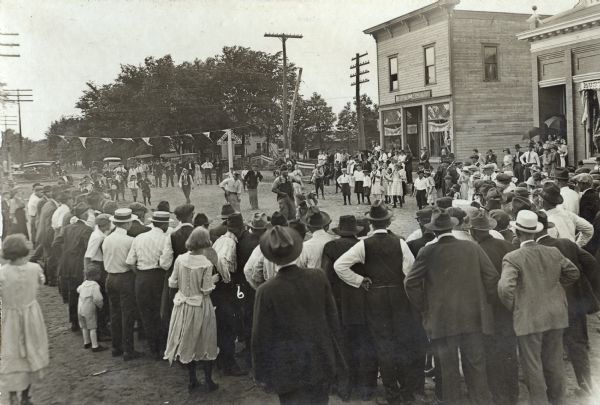 A laughing and smiling crowd of people surrounds a sack race at the Harvest Festival. Across the street is a storefront with a sign for DeLaval Cream Separators, and also the Hustler State Bank. Cars are parked in the street in the background behind the starting line.
