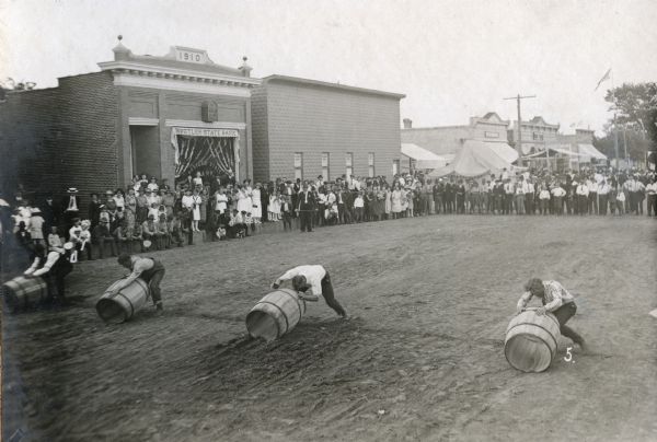 Four men struggle to move barrels down the dirt main street of Hustler. It looks like a close race. The large crowd watches attentively. Across the street is the Hustler State Bank.