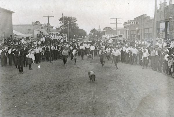View down unpaved Main Street of a group of men racing after a pig during the Hustler Harvest Festival. On the right is the Nash Service Station and the M.O. Rider Garage.