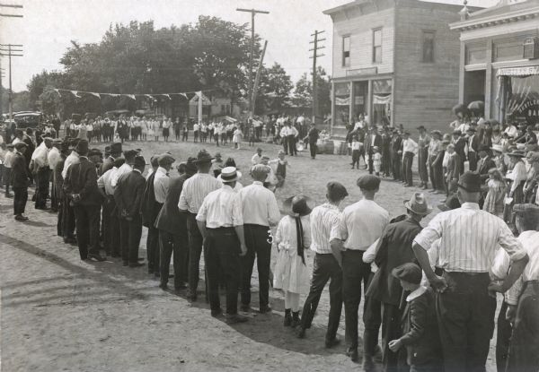 A group of young boys races down the street as a large crowd watches. The Hustler State Bank is across the street on the right. A group of people stand in a line at the starting line near the railroad crossing.