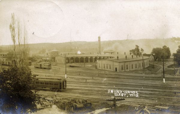 Elevated view of the roundhouse for the Chicago North Western Railway, one of two Elroy roundhouses during the railroad boom. This roundhouse has 20 stalls for train cars, and there are two cars in the stalls. Outside the roundhouse are several single cars, as well as a train. In the foreground is a single passenger car and several pieces of railroad equipment. In the far background are tree-lined hills.<p>The railway boom slowed in the early 1920s and ended in the 1960s. The railroad tracks were removed and converted to bike trails in the 1990s.