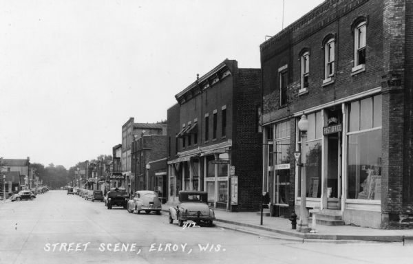 View down Main Street. The wide street has cars and trucks parked along the curbs on both sides. The Elroy Post Office is on the corner on the right. Further down is a doctors office, a shoe store, a drug store, a barber shop, a bakery and a meat market. A delivery truck is sitting outside the bakery in the street with a back door open.