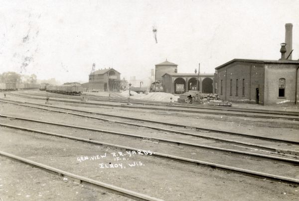 View of the Chicago North Western Railroad Roundhouse in Elroy. There are several train cars sitting on multiple sets of railroad tracks on the left. There are two cars in the roundhouse stalls.
