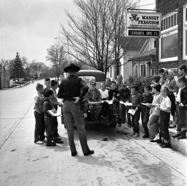 Sterling Standiford, a State Trooper, talking to a group of local children on bicycle safety during National Bicycle Week. The activities took place outside of Sherman Hardware and Massey Ferguson Farm Equipment. Some of the children are sitting in back of a pickup truck.
