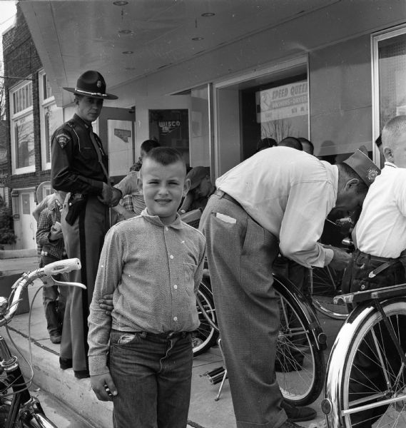 A boy taking part in a National Bicycle Week Celebration stands near a bicycle. Sterling Standiford, a State Trooper who  was present to explain proper operation of bicycles on streets and highways, is standing behind him, near members of the Shaker-Callaway Post of the VFW who were on hand to place strips of Scotch Lite on the bicycles. The activities took place outside of Sherman Hardware.
