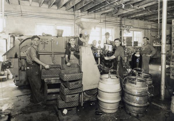 The interior of the bottling house at Hillsboro Brewery. The bottling equipment is numbered 1 through 5 and the following description is included with the photograph: "1. Alkale Bottle Washer; 2. Crowner; 3. Twelve Spout Rotary Bottle Filler; 4. Keg Beer Supply-31 gallons each; 5. Carbonated Gas." The following names also appear in the description (presumably the names of the workers in the photograph): "Abbey; Charley; Johnson; Fibber; Rinkey; French."