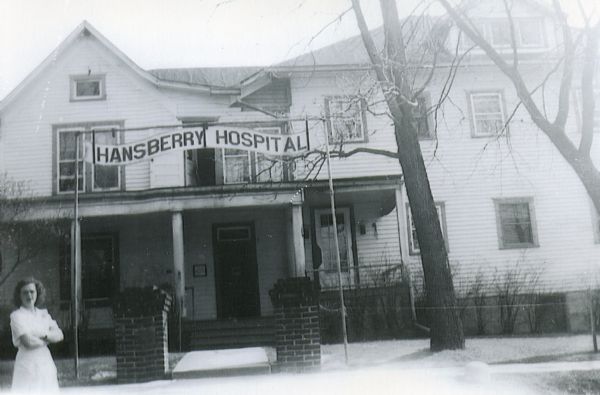 A woman (possibly a nurse) standing on the sidewalk in front of the sign at the entrance to Hansberry Hospital, Hillsburo's first hospital. It was established by Dr. P.H. Hansberrry in 1914.