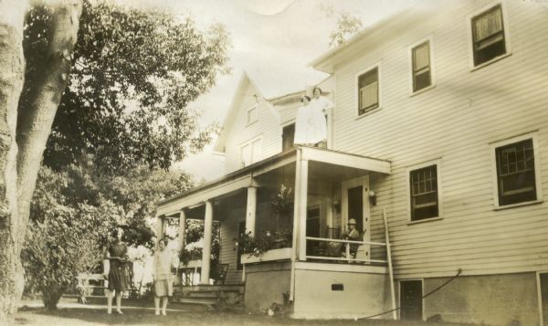 Three-quarter view from yard of Hansberry Hospital, Hillsburo's first hospital which was established by P.H. Hansberry in 1914. Two women stand on the left near the sidewalk leading to the porch entrance. A woman sits in a rocking chair on the columned porch, which has flower boxes along the front.