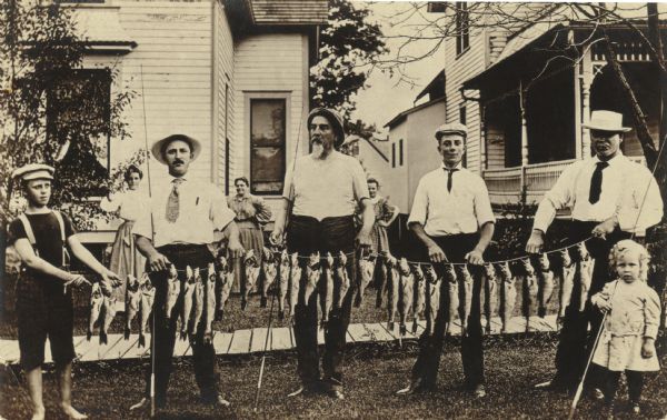 Group portrait of men and boys standing in front of two houses all holding a long stringer of fish. Three women are standing in the yard in the background. Front row, left to right: Conrad L. Kolb, Edward John Hammer, Augustus Weinstein, Conrad Hammer, Grutav Kolb, Russell Kolb. Back Row, left to right: Lina Marsh Kolb, Margret Weinstein Kolb and Margaurite Kolb Horkan.