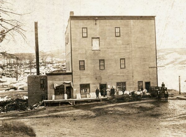 View from dirt road of a group of people (possibly employees) standing around the loading dock of the Hillsboro Mill. There is a steep, snow-covered hill in the background. In 1865, locals Edward Klopfleisch, Otto Hammer, John Mollinger, and Ambrose Armbruster built the first grist mill in Hillsboro. These men and their families operated the mill until it was sold to the Vernon County Milling Company in 1908.