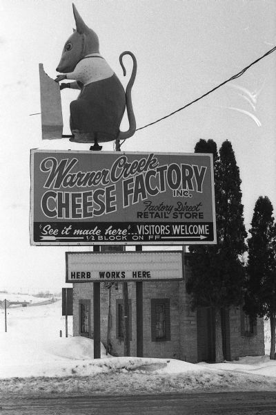 Warner Creek Cheese billboard, including a 3D sculpture of a mouse with cheese, in front of a small, brick building. Located off the State Highway near Hillsboro. The Warner Creek Cheese Factory moved to Hillsboro in 1971. In 1985, the factory was sold to a different owner, however, the sign remained to attract customers.