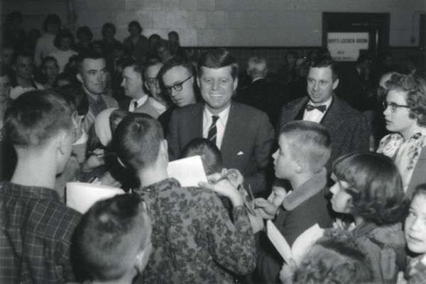 John F. Kennedy surrounded by Hillsboro High School students after a campaign speech. Kennedy, then a Senator, spoke at the Hillsboro High School Auditorium during his Presidential Primary Campaign in Wisconsin. Photographs appeared in the <i>Hillsboro Sentry Enterprise</i>.