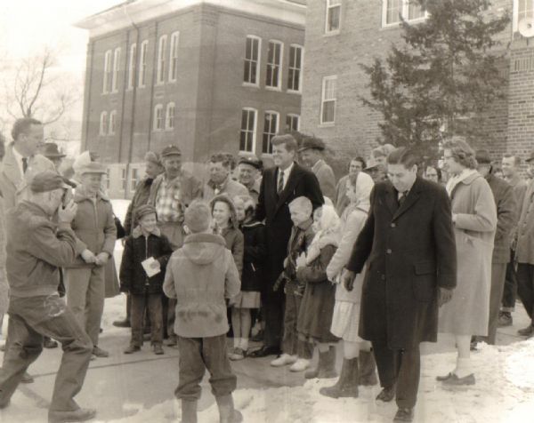 John F. Kennedy outdoors with a group of Hillsboro students and residents after a campaign speech. A man on the left is taking a group photo with a camera. Hillsboro High School is in the background. Kennedy, then a Senator, spoke at the Hillsboro High School Auditorium during his Presidential Primary Campaign in Wisconsin. Photographs appeared in the <i>Hillsboro Sentry Enterprise</i>.