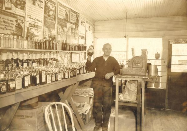 Interior view of Hofmeister's Liquor Store. John Hofmeister is standing and holding up a bottle next to the cash register.
