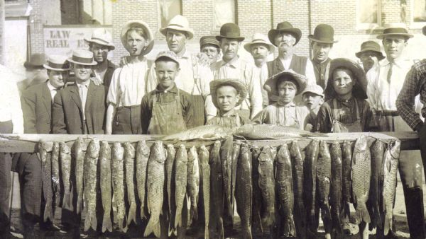 Group portrait of five local youth showing off a record breaking catch of thirty large Carp hanging on a stringer. Town elders, all men, pose in the background.