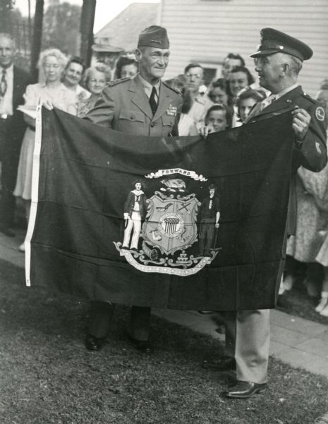 Vice Admiral Marc Mitscher, wearing his uniform, is being presented with a flag of the State of Wisconsin by an unidentified man, who is also wearing a uniform. A crowd of people stand in the background. It was the first time in ten years that the military commander had visited his birth place. The town honored Mitscher with a party and parade. Local and state newspapers covered the event.