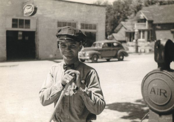 Rudy Lein, owner of the Standard Oil Gas Station, standing next to an air pump outside of the station. He wears a Standard Service cap and a bowtie. Behind him an automobile is parked in front of what appears to be a garage.
