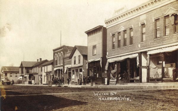 View across unpaved street of people, horses and wagons, and businesses on one side of Water Avenue, the heart of Hillsboro's downtown and business district.