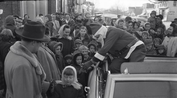 A man dressed as Santa Claus leans down from an open car to hand sacks of candy to a crowd of warmly-dressed children and parents. Santa's visit took place in front of Farmers State Bank on Water Avenue. The Hillsboro Lions Club organized the event.