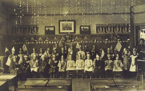 Group portrait of class of young students holding American flags posed at the front of a classroom. Their teacher is standing on the far right, and desks are in the foreground. The students are wearing paper hats styled after those worn by soldiers in the Revolutionary War. The chalkboard in the background is decorated with hatchets, snowflakes, and a portrait of George Washington. A cloth with a pattern of Christmas stockings hangs above the chalkboard, and strings of popcorn hang from the ceiling. The holiday that the students are celebrating is not known, however, considering the Christmas decorations, it is likely that the students are commemorating Washington's crossing of the Delaware, which occurred from December 25 to December 26, 1776.