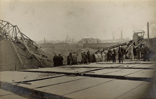 View across what may be a metal roof towards a crowd of men looking at a collapsed structure of steel girders. The men are dressed in winter coats and hats. In the distance is a large pile of what may be coal, and a sign on an industrial building reads: "Coal / Wood." In the far background is the skyline of Milwaukee, with steeples and chimneys.