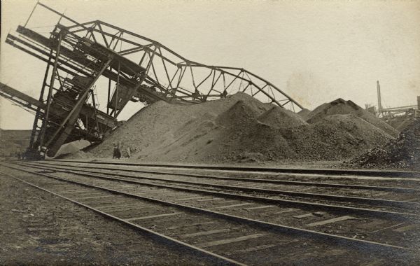 View across railroads tracks towards men walking around and examining a large industrial structure that has collapsed over a coal pile.
