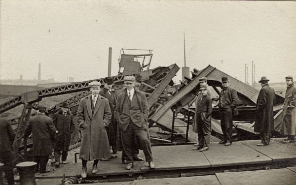 Group portrait of men wearing winter coats and hats. They are standing on a metal roof, examining a large collapsed industrial structure which is behind them.