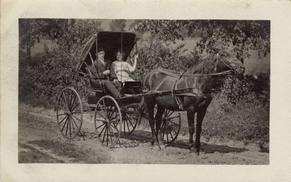 William Unglaube is in a two-seater buggy with one of his sisters. The buggy is pulled by one horse on a dirt road.
