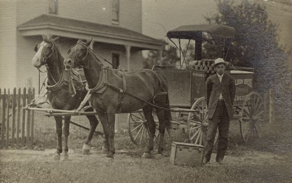 A traveling salesman, dressed in a suit and light hat, stands beside his carriage. A case of perfume and remedy wares sits near his feet. The small carriage is pulled by two horses and is stopped in front of a house.