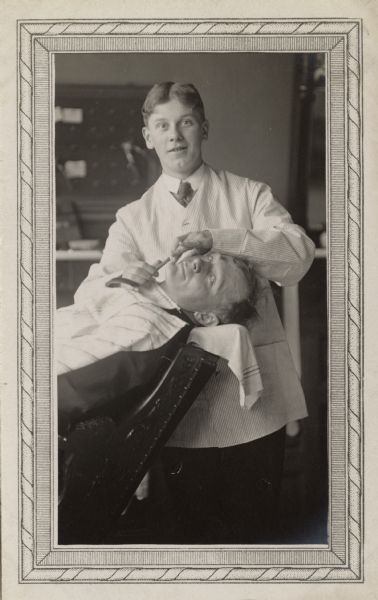 A barber is smiling for the camera, while the customer resting back in the chair in front of him is looking on with a sidelong look.