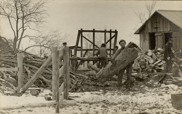 A group of men are working on sawing logs into smaller pieces. In the foreground, an unidentified man is holding a large piece of tree trunk.