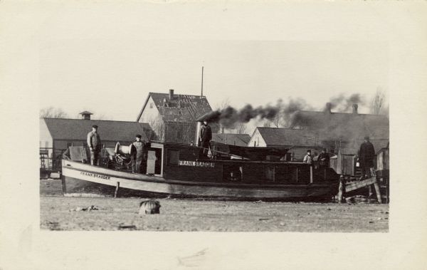The <i>Frank Braeger</i> is moored on the shore of Jones Island with steam issuing from the pipe, with the crew posing on the boat. In the background a man is roofing a house. The <i>Frank Braeger</i> tug boat was used for fishing from 1906 until 1931 and ran on Lake Michigan. Frank Braeger and his family lived on Jones Island.