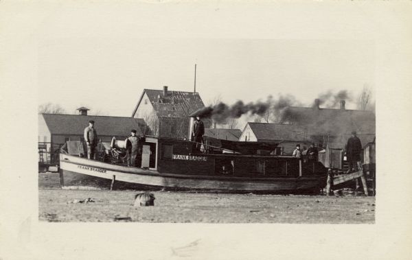 The <i>Frank Braeger</i> is moored on the shore of Jones Island with steam issuing from the pipe, with the crew posing on the boat. In the background a man is roofing a house.

The <i>Frank Braeger</i> tug boat was used for fishing from 1906 until 1931 and ran on Lake Michigan. Frank Braeger and his family lived on Jones Island.