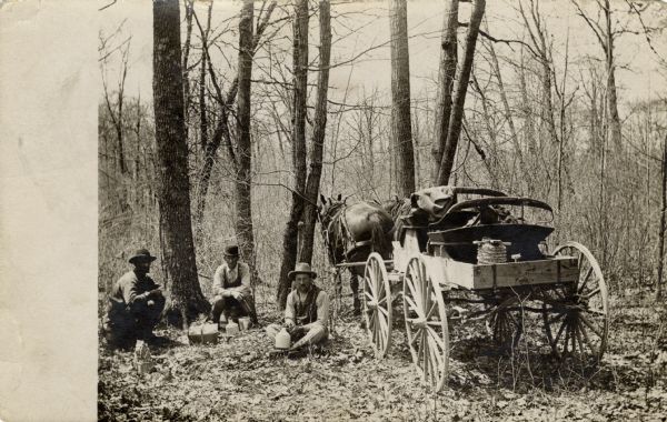 Three men sitting on the ground next to a horse-drawn wagon are having lunch. Two of them have jugs; one is holding a cup. All three are wearing work clothes and hats. The wagon has a roll of barbed wire in the back. The postcard is from Ida Unglaube (William Fred) Fiebelkorn to her sister Louise (George) Koegel.