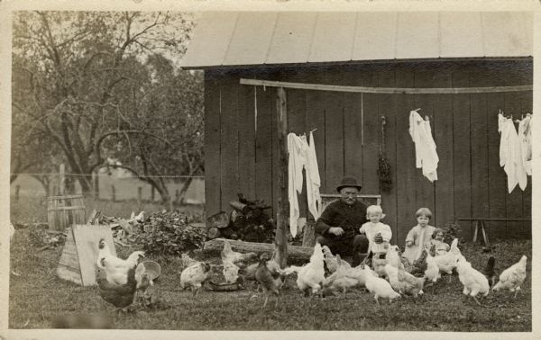 A smiling grandfather has his arm around a toddler granddaughter. A toddler boy is standing next to them. They are surrounded by chickens, and behind them clothes are hanging on a clothesline. In the background is a shed or barn.