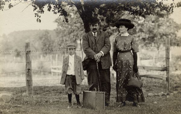 Group portrait of George, Louise, and Milton, their son, standing in front of a fence. They are traveling with one suitcase and a full handbag. All three are wearing hats.
