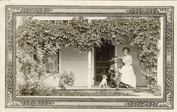 A young couple poses on their vine bower framed porch, with a pet Jack Russell terrier. The man is sitting in a chair in front of the screen door holding a hat, and the woman is sitting on the arm of the chair. She is wearing a long dress and two-toned shoes tied with a bow. There are lace curtains on the window.