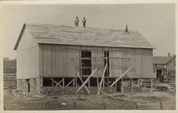 Four men pause in their work of building a new, large barn. They are all wearing hats for protection from the sun.